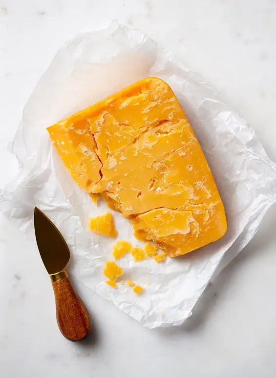 A piece of orange cheese on paper beside a cheese knife