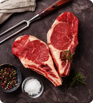 Two raw steaks beside salt, peppercorns and a carving fork
