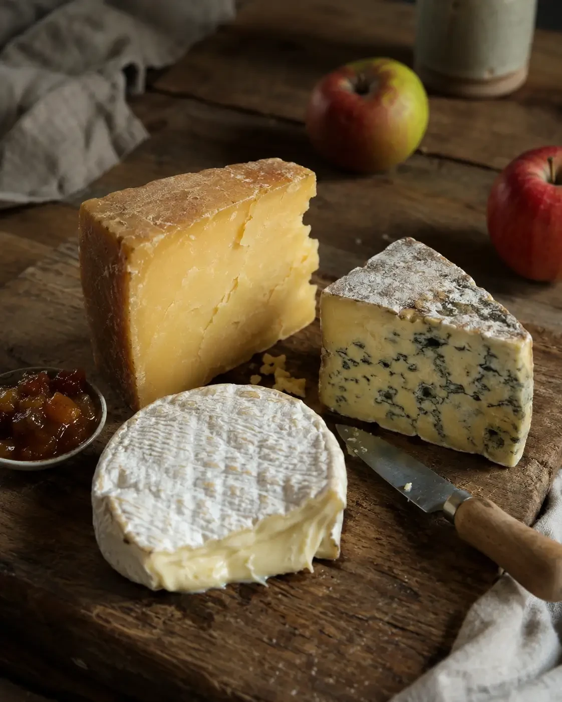 Soft, blue and hard cheeses on a board beside apples and chutney