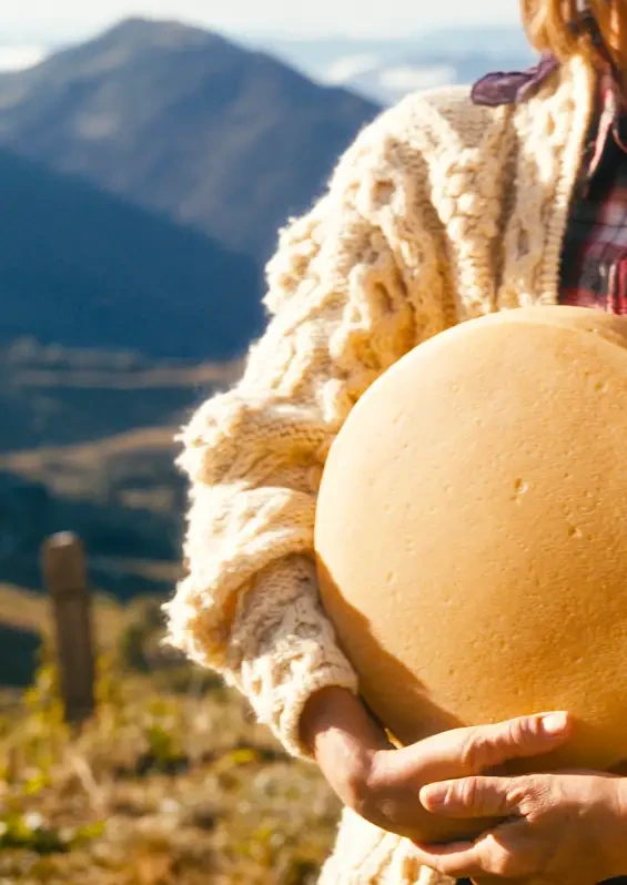A person holding a large cheese wheel outdoors with mountains behind
