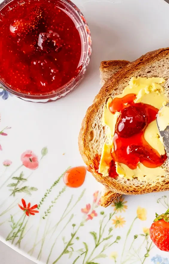 Butter and red jam on a slice of toast beside an open jam jar