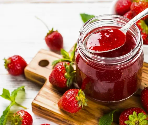 An open jar of strawberry jam with a spoon and fresh strawberries