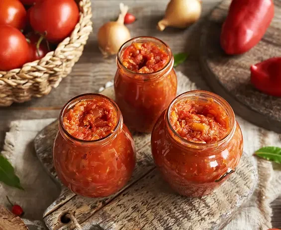 Three open jars of red vegetable relish beside tomatoes and peppers