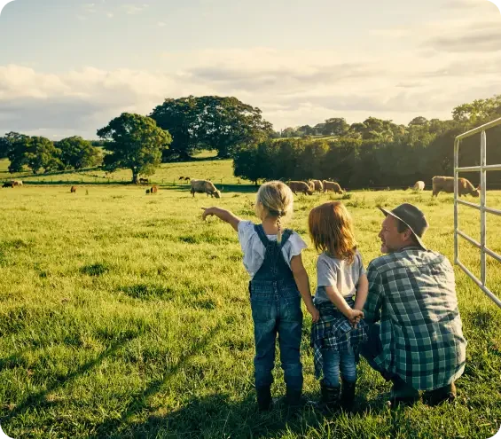 Three children in countryside clothing pointing at grazing cows in a sunny British pasture with trees