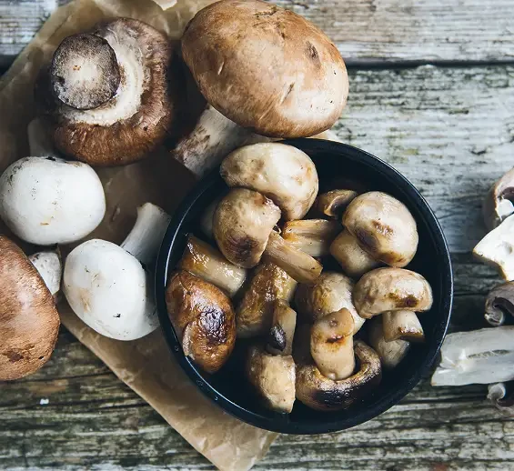 Fresh mushrooms beside a pan of cooked mushrooms on a wooden table