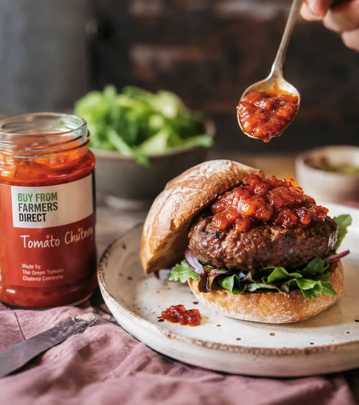 A spoon adding tomato chutney to a burger beside a chutney jar
