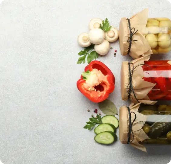 Jars of preserved vegetables beside mushrooms, peppers and cucumber