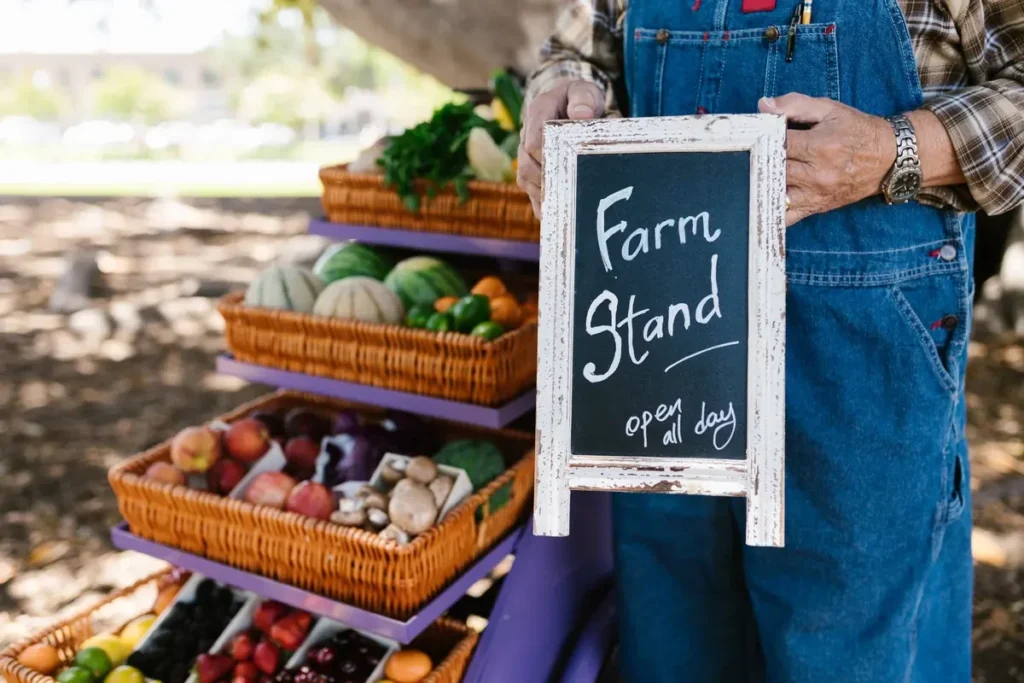 Farmer in overalls holding 'Farm Stand open all day' sign beside baskets of fresh vegetables